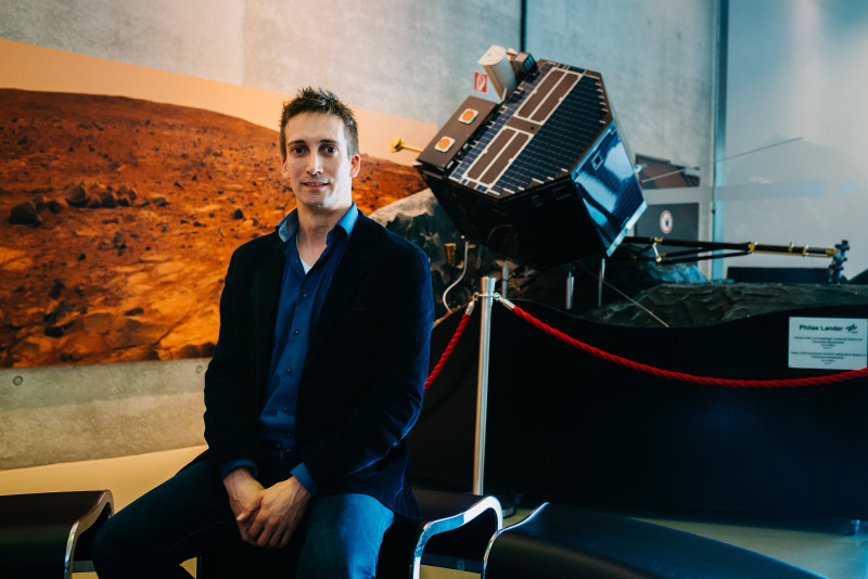 Ein junger Mann in blauem Hemd sitzt im Vordergrund, im Hintergrund ist Demomaterial zu Landetechnologien auf fremden Planeten zu sehen