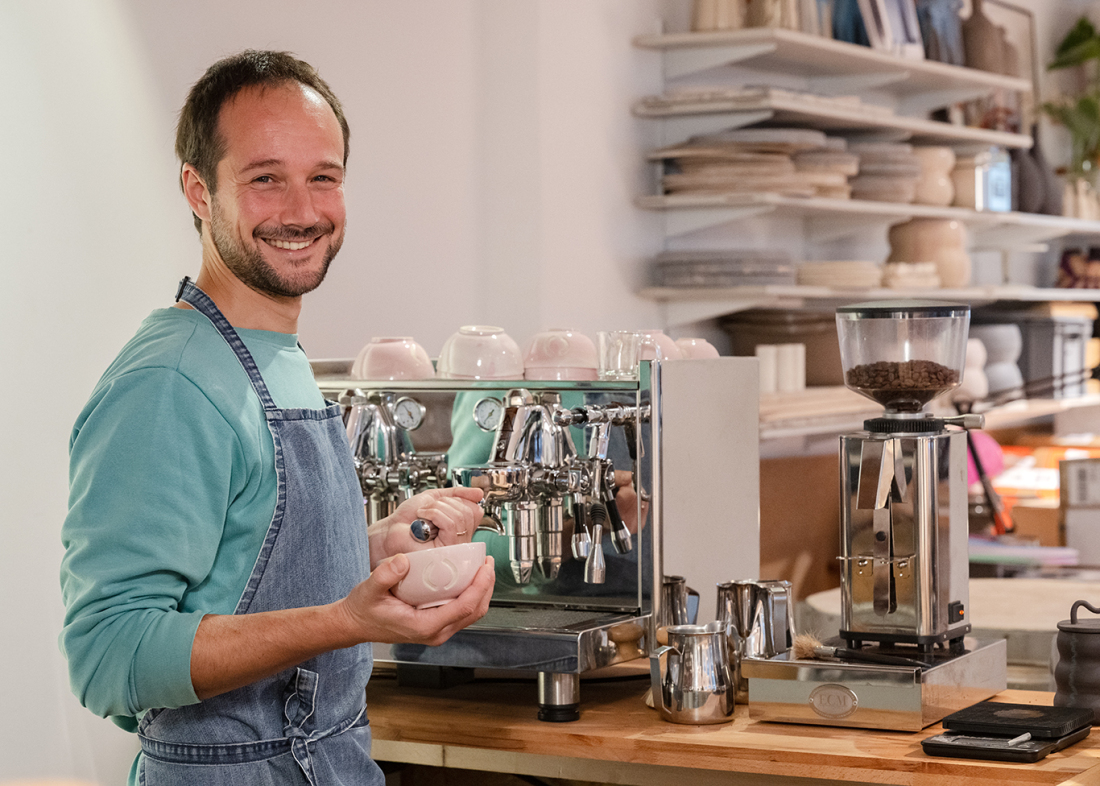 Ein Mann mit Schürze aus blauem Jeansstoff steht lächelnd an einer großen Siebträgermaschine. In der Hand hält er eine Kaffeetasse, im Hintergrund hängen die Regale einer Keramikwerkstatt.