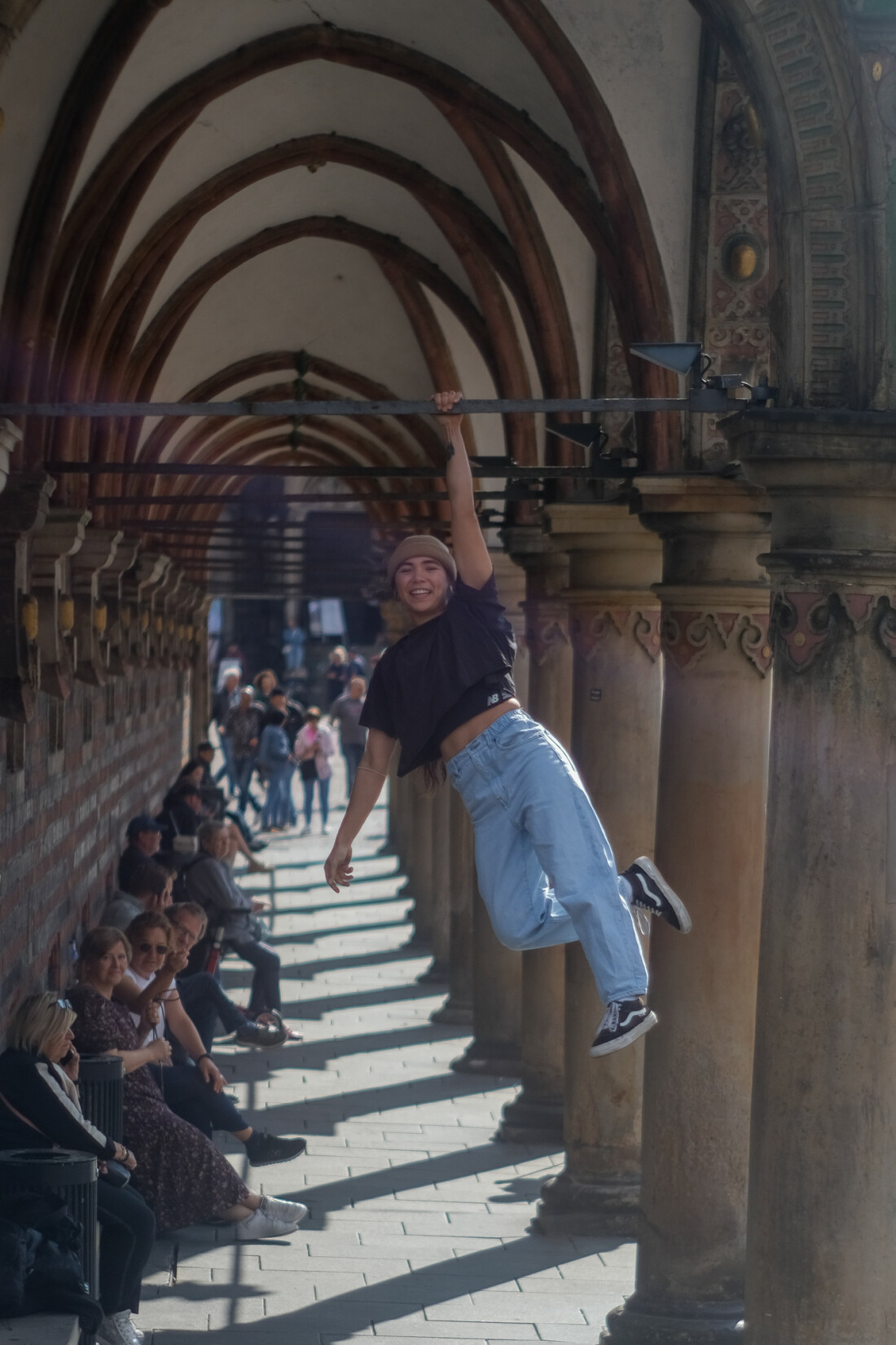 Eine junge Frau in Jeans und Shirt hängt lächelnd an einer Metallstrebe in ungefähr drei Metern Höhe. Diese gehört zu den Arkaden des Bremer Rathauses.