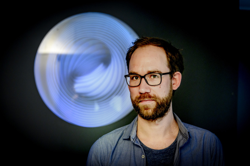 Ein Mann mit kurzen braunen Haaren, schwarzer Brille und Bart blickt in die Kamera. Hinter ihm: eine dunkle Wand und ein kreisförmiges, hell leuchtendes Objekt.